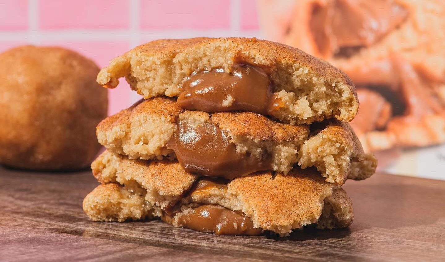 A stack of three La Gringuita gourmet cookies is broken in half, revealing a gooey chocolate filling inside. A round dough ball and a blurred background complete the mouthwatering scene.