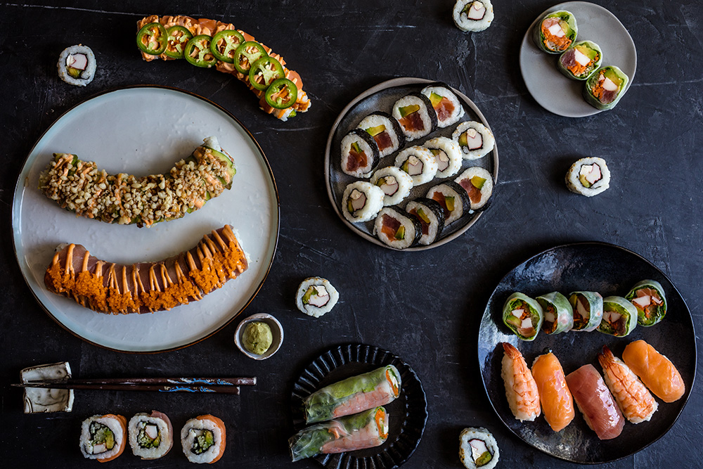A variety of sushi rolls, nigiri, and sashimi made with fresh seafood are arranged on dark plates and a dark tabletop, with soy sauce, wasabi, pickled ginger, and chopsticks also visible.