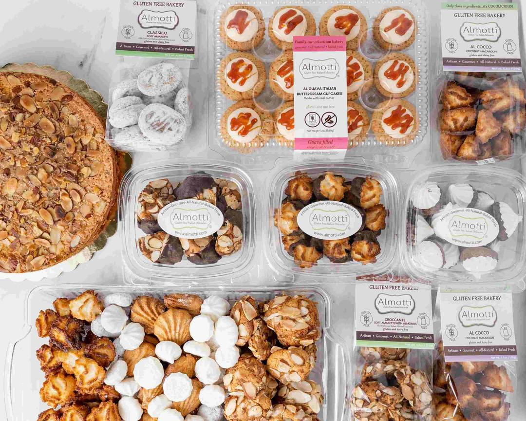 An assortment of Almotti gluten-free baked goods, featuring Italian delicacies like almond cake, cookies, meringues, biscotti, and cupcakes in clear containers with Almottti Bakery labels, displayed on a white surface.