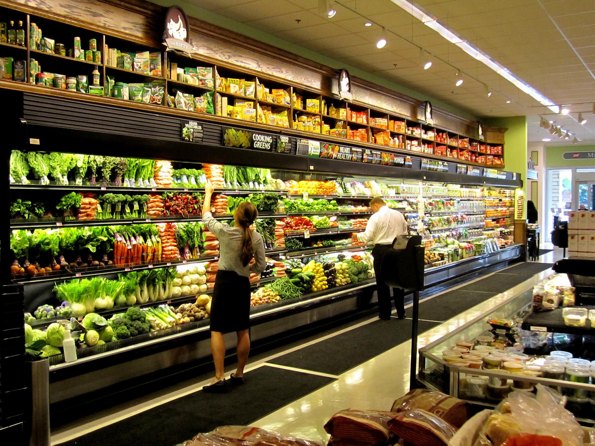 A woman and a man select fresh produce from a well-stocked grocery store aisle filled with various fruits, vegetables, and leafy greens under bright lighting.