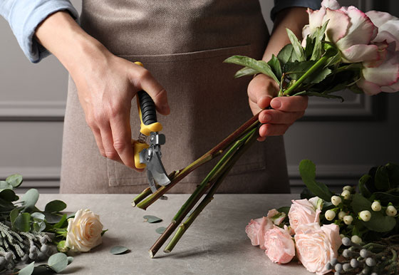 A person wearing an apron is using pruning shears to trim the stems of a bouquet of roses and greenery on a gray table, with more flowers and leaves scattered nearby.