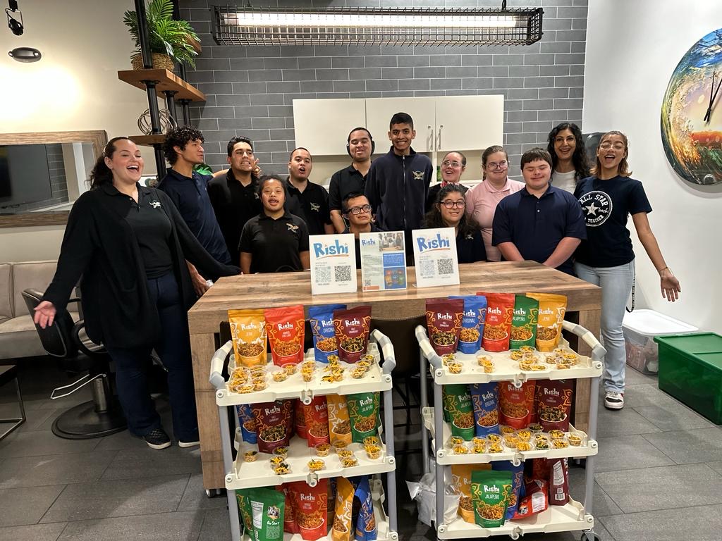 A group of people, some in matching shirts, stand smiling behind a wooden counter with two carts filled with colorful Rishi Snacks bags and clear tubs, showcasing community empowerment in a modern indoor setting.