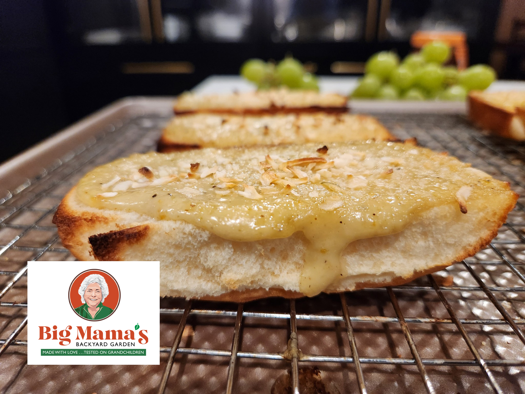 A slice of toasted garlic bread with melted cheese and herbs sits on a wire rack. In the background are green grapes. The Big Mamas Backyard Garden logo is displayed in the lower left corner, promising the world’s best garlic bread experience.