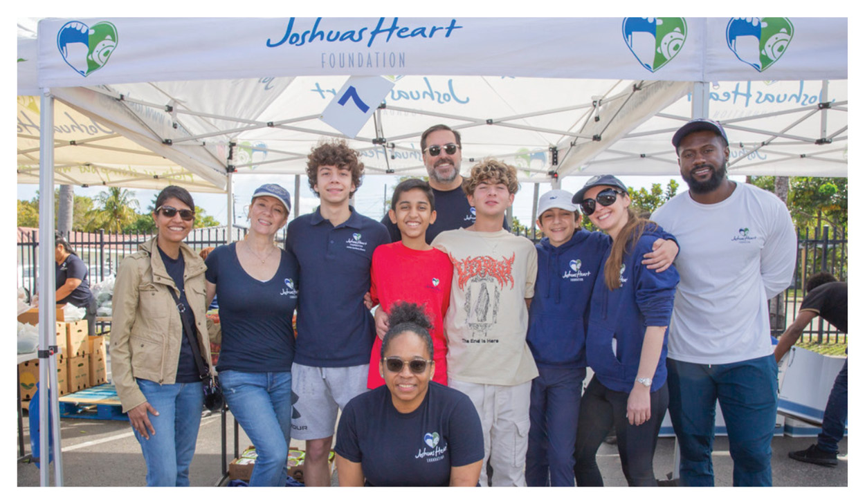 A group of people, both teens and adults, smile together under a tent with Joshua’s Heart Foundation written on it. They are outdoors on a sunny day, wearing casual clothes and foundation-branded shirts, supporting local food banks through donation.