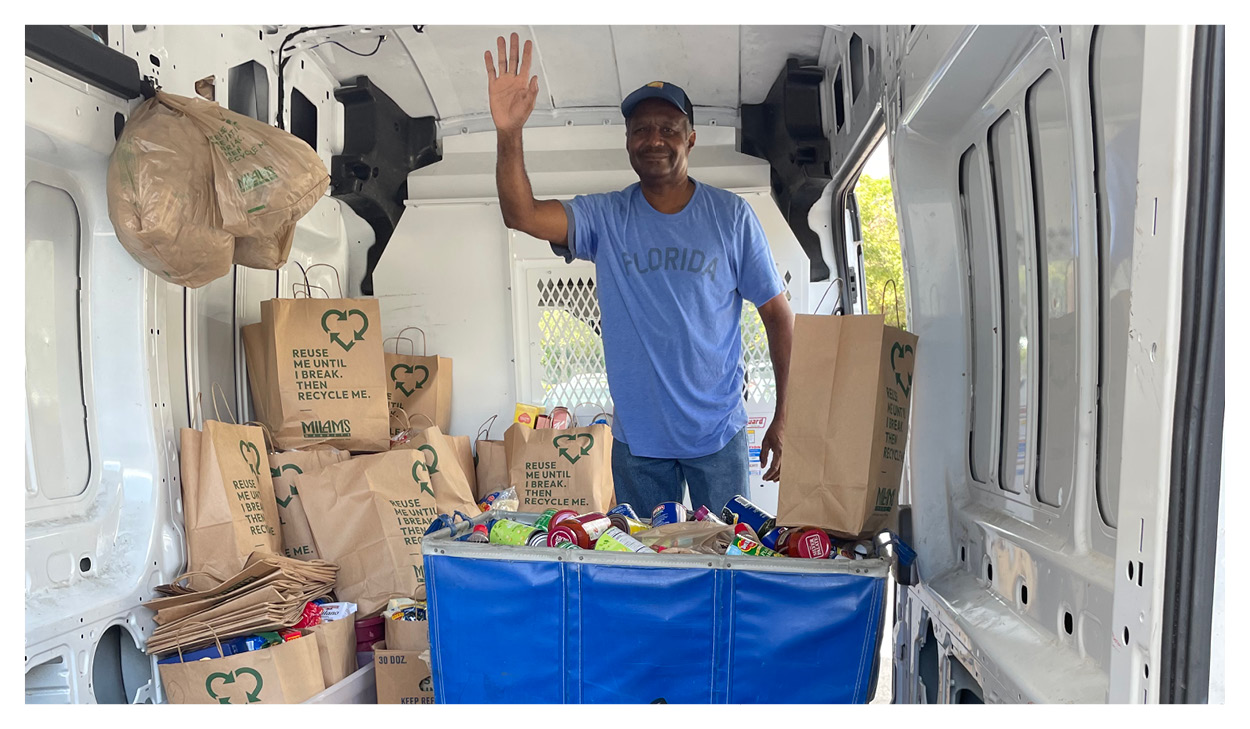A smiling man wearing a blue Florida shirt stands inside a van, waving. The van is filled with paper grocery bags and canned goods, showing support for local food banks through a recent fundraising effort.