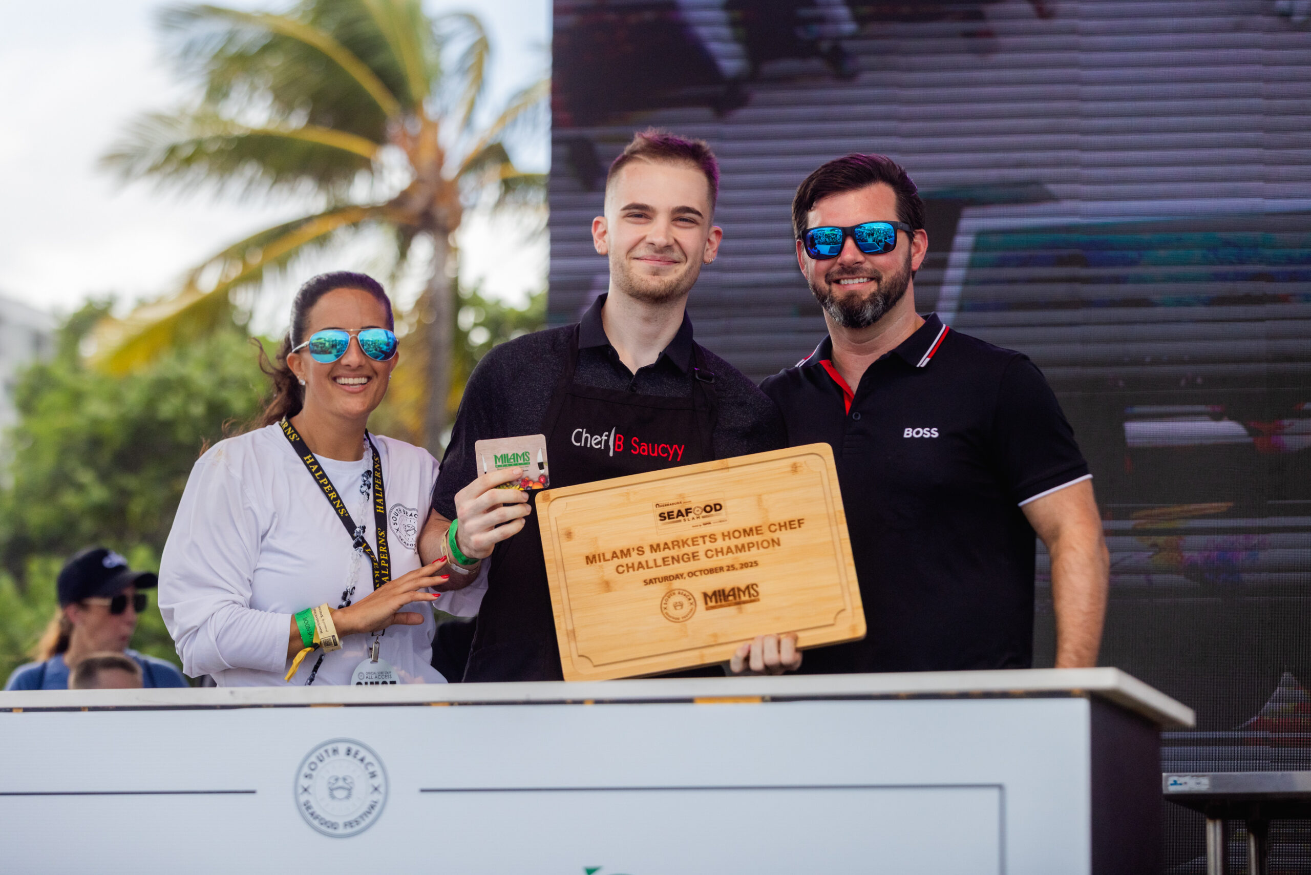Three people stand together outdoors, smiling at the camera. The person in the middle holds a wooden award plaque, perhaps celebrating victory in a Tacos competition. Palm trees and a large screen hint at a lively festival atmosphere.