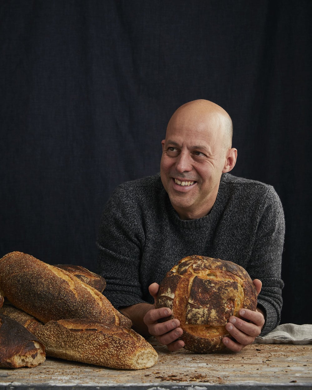 A smiling person with a shaved head and a grey sweater holds a round loaf of rustic bread, with several other loaves from Sullivan St. Bakery stacked on a floured surface in front of a dark background.