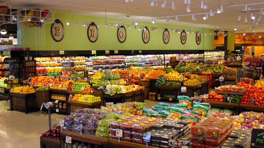 A bright produce section in a grocery store filled with various colorful fruits and vegetables neatly arranged on displays, with a large PRODUCE sign on the green wall above.