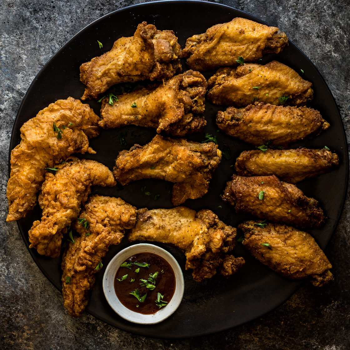 A black plate filled with crispy fried chicken wings garnished with herbs, arranged in a circle around a small bowl of dark dipping sauce, set on a textured dark surface—perfect for those who want to order online and enjoy at home.
