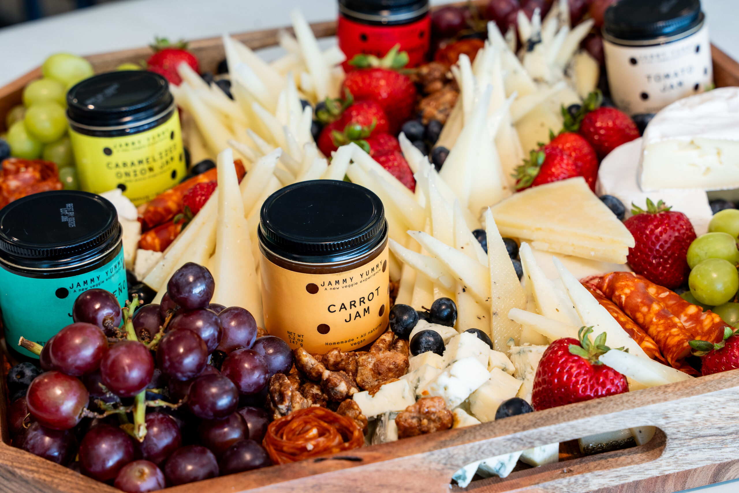 A wooden tray filled with assorted cheeses, carrot jam jars, strawberries, grapes, walnuts, blueberries, green grapes, and sliced cured meats—perfect for exploring unique cheese pairings on a beautifully arranged charcuterie board.