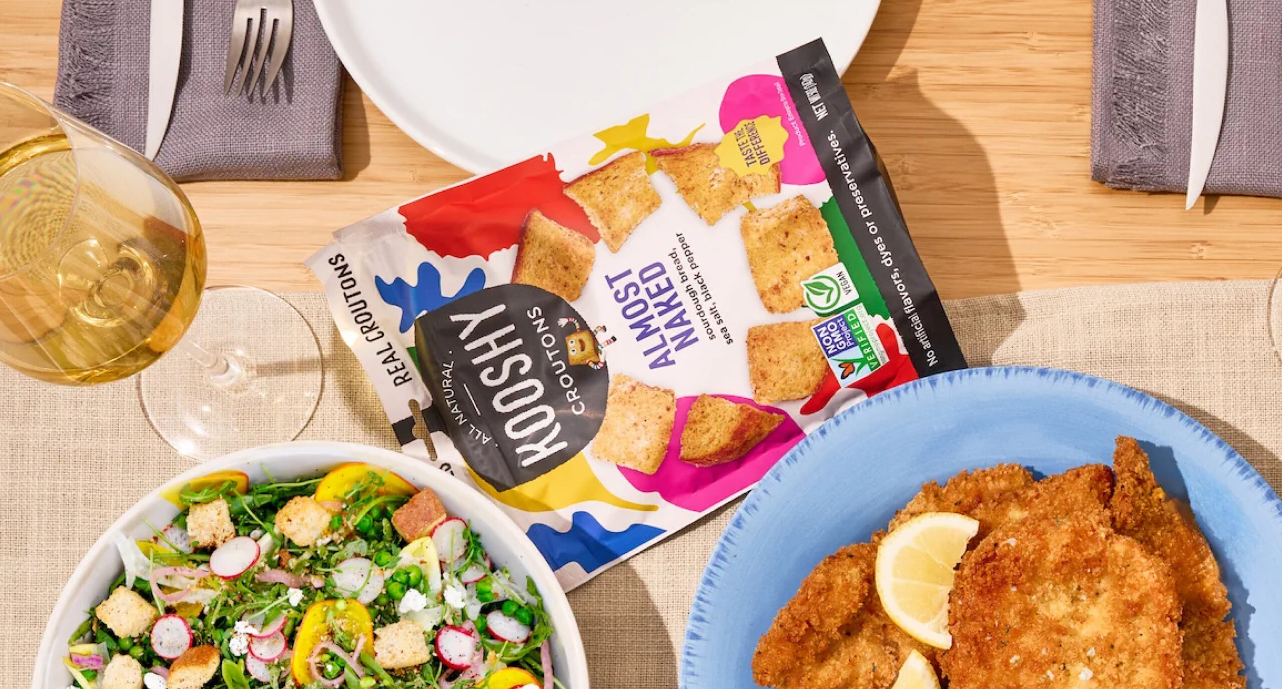 A table set for a meal with a plate of breaded cutlets and lemon slices, a salad with radishes and greens, a glass of white wine, and a bag of Kooshy Croutons from the New Arrivals collection.