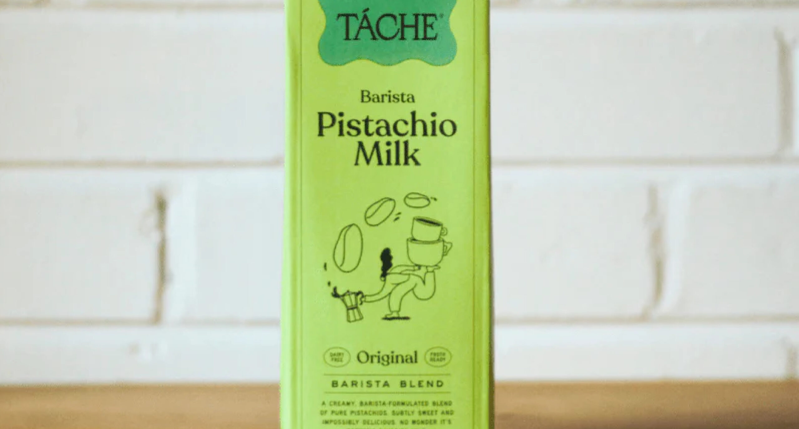 A green carton of Tache Barista Pistachio Milk, part of our New Arrivals, features an illustration of a cup holding a spoon and pistachios, set against a white brick wall background.