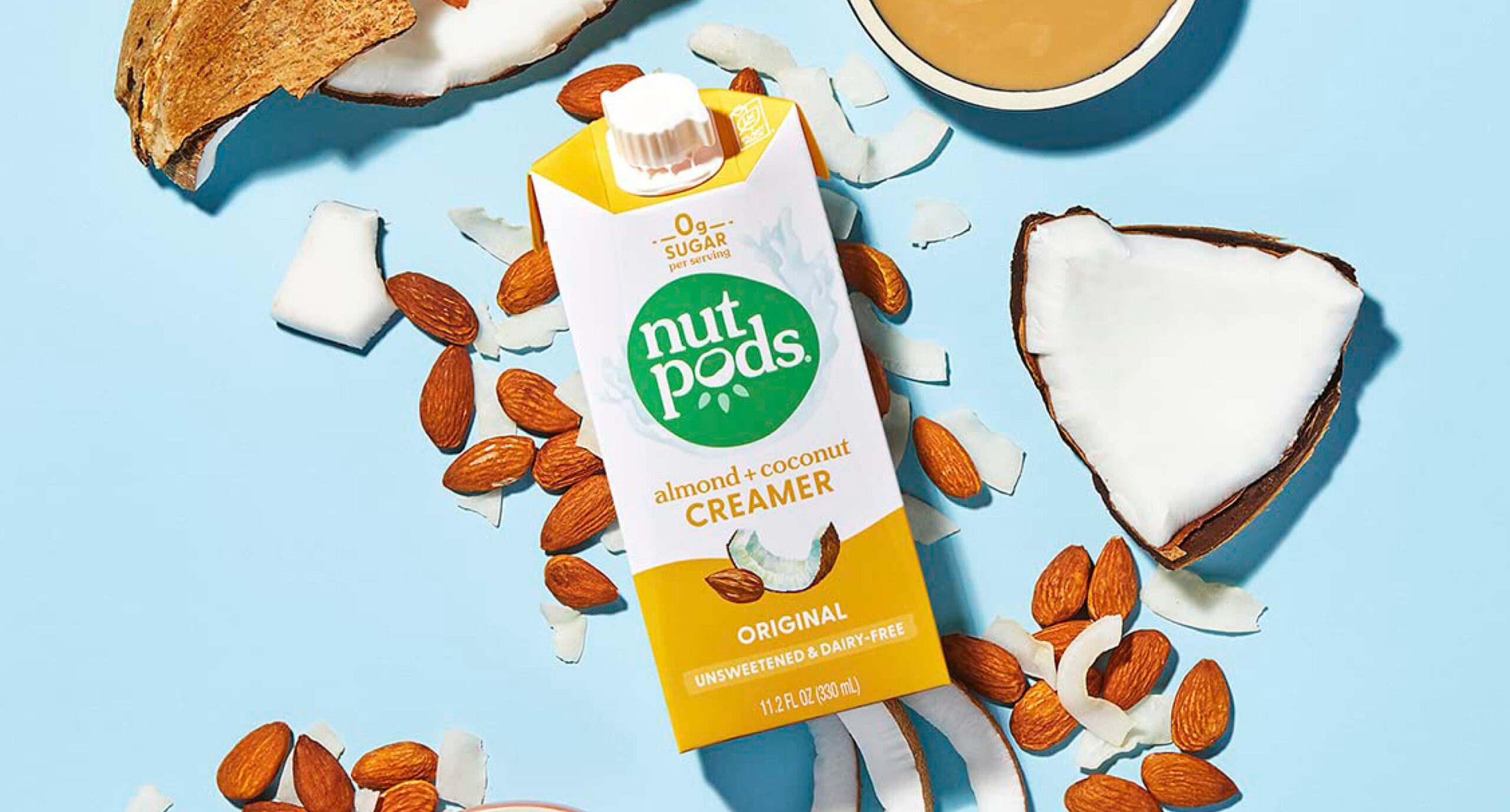 A carton of Nutpods almond + coconut creamer, a New Arrivals favorite, is surrounded by whole almonds, coconut pieces, and a cup of coffee on a light blue background. The unsweetened creamer is labeled dairy-free.