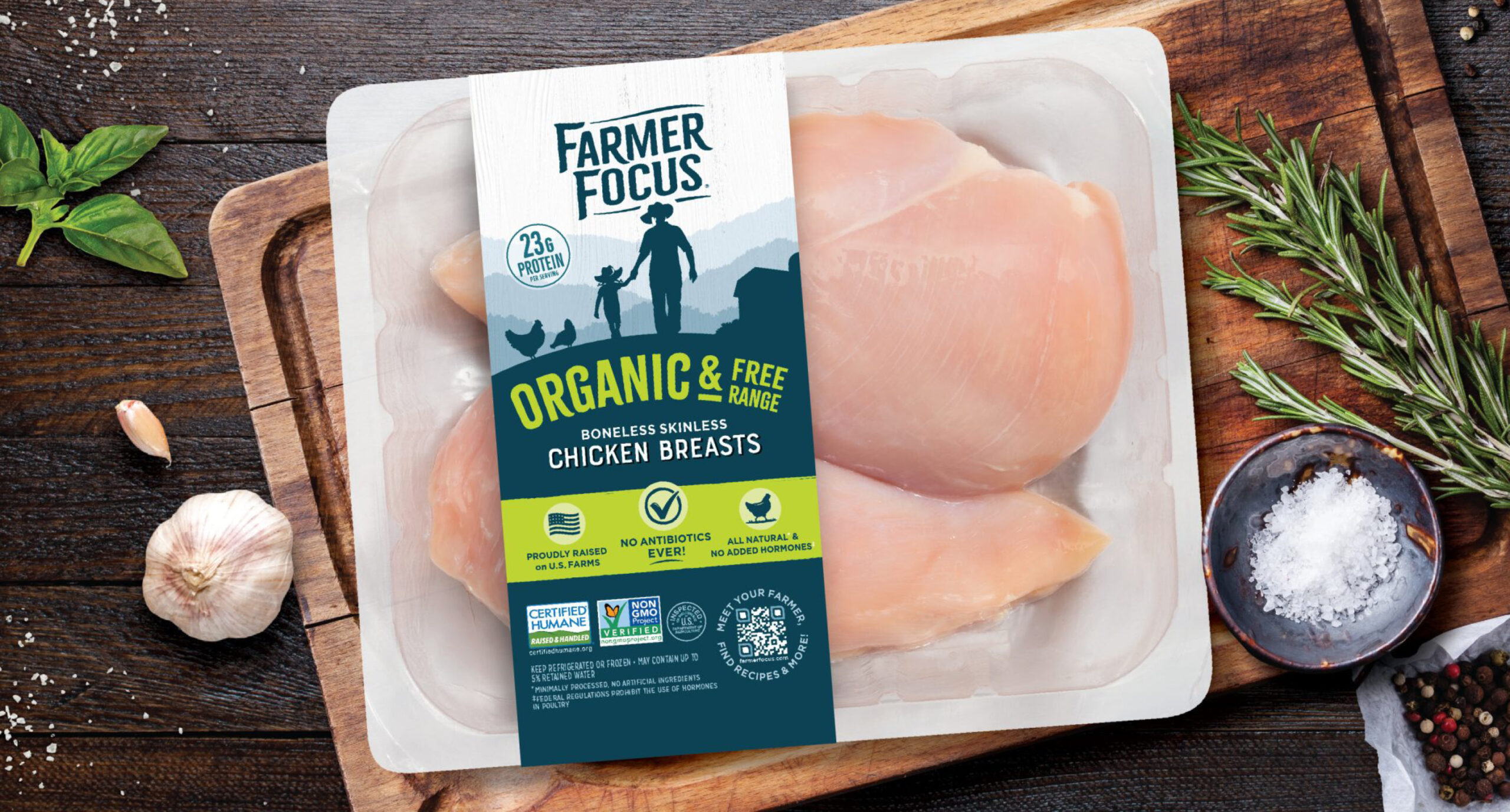 A package of Farmer Focus organic, free-range boneless skinless chicken breasts sits on a rustic wooden table with fresh herbs, garlic, black peppercorns, and lime. The label highlights protein content and humane certification—perfect for New Arrivals to your kitchen.