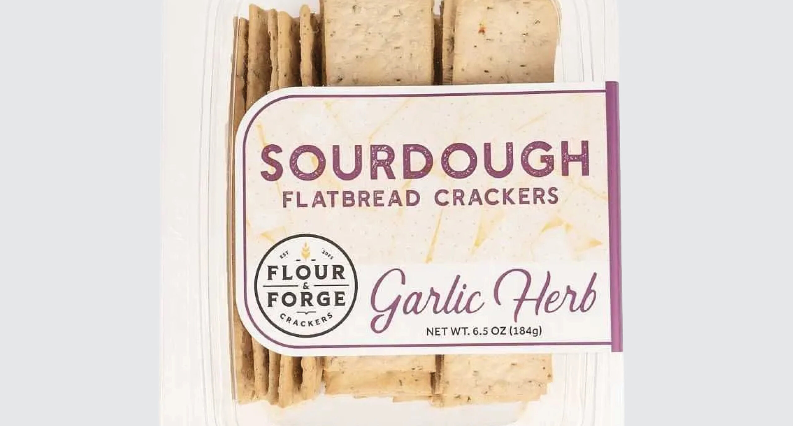 A clear plastic container of Flour & Forge Sourdough Flatbread Crackers in Garlic Herb flavor, part of our New Arrivals. The crispy crackers are visible through the packaging, and the label features a clean white and purple design.
