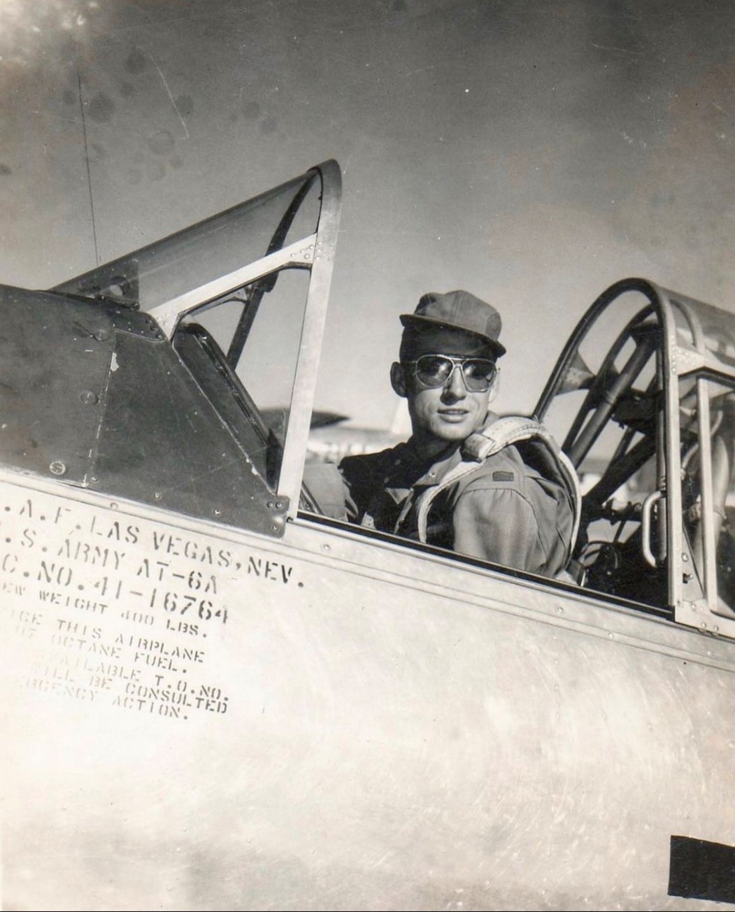 A pilot wearing sunglasses and a cap sits in the cockpit of a military aircraft, looking toward the camera. The side of the plane shows identification markings and text. The sky is clear in the background.