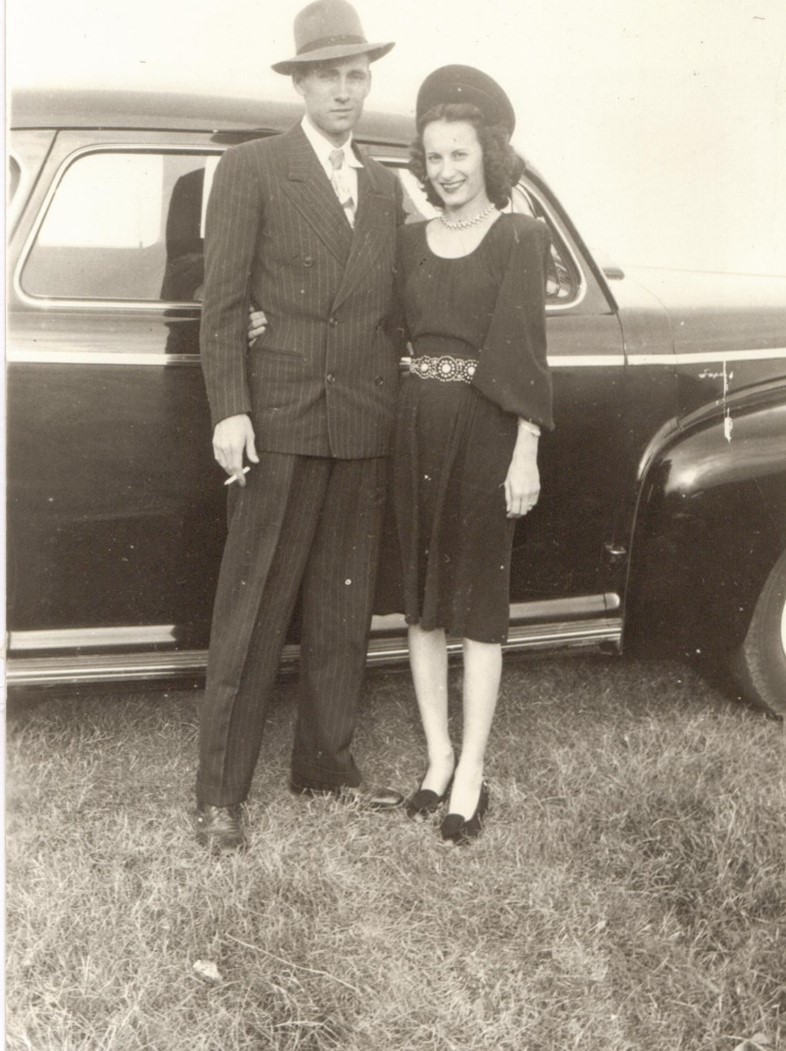A man in a pinstripe suit and fedora stands with his arm around a woman in a dark dress and hat, both smiling, in front of a vintage car parked on grass in a black-and-white photo.