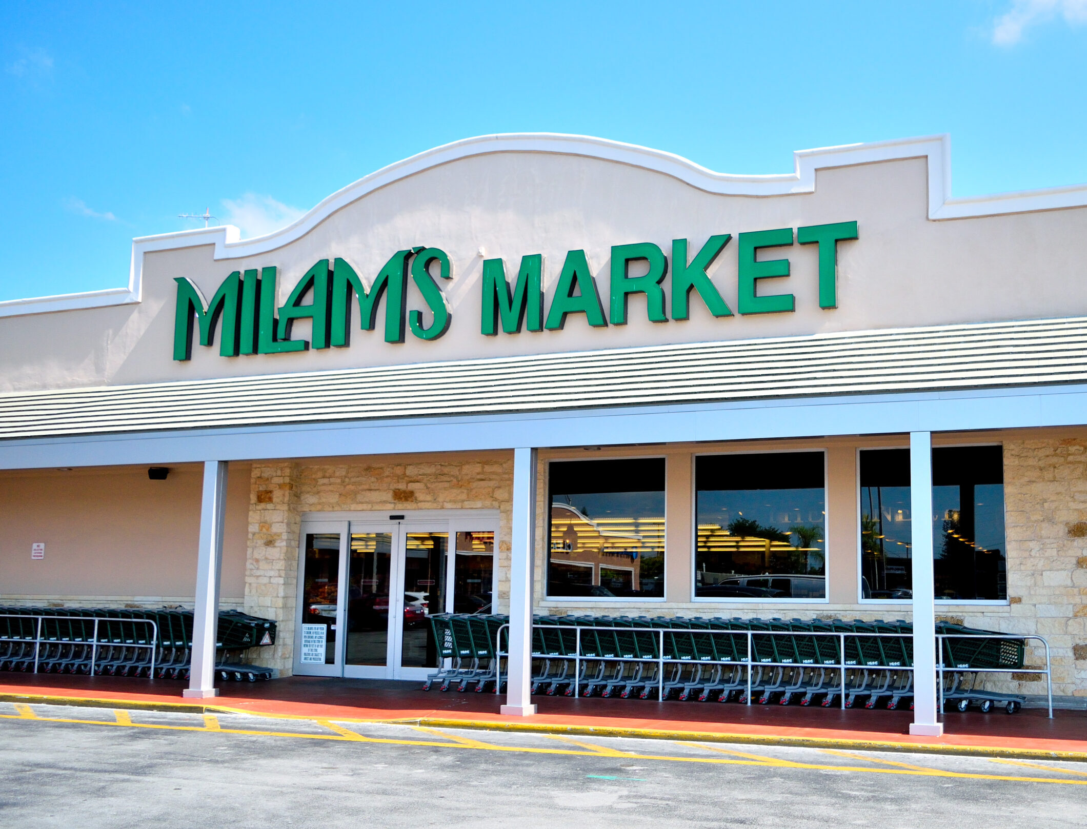 The exterior of Milams Market in Coral Gables West features a large green sign, glass entrance doors, and rows of shopping carts outside under a sunny blue sky.