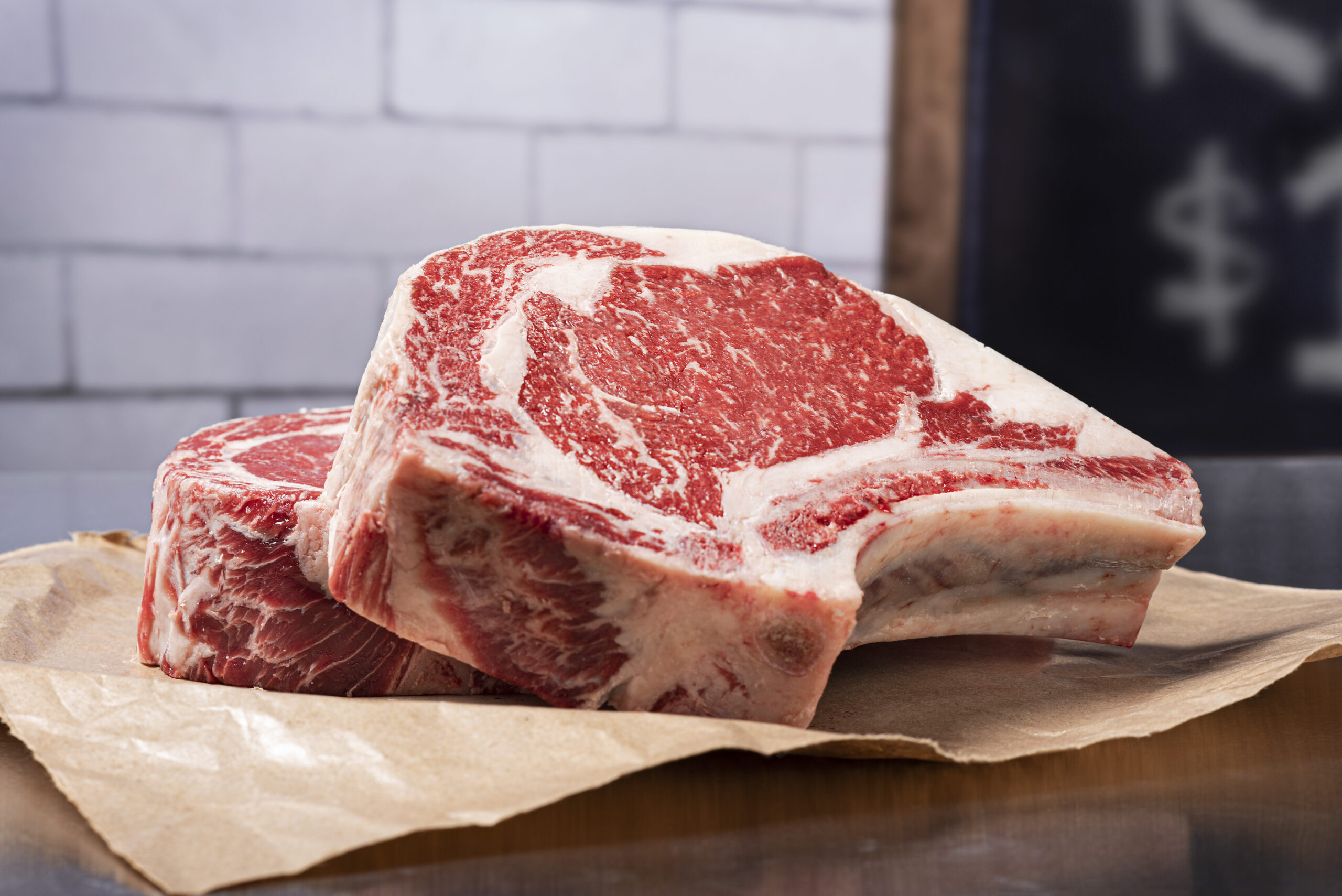 Two raw, marbled ribeye steaks sit stacked on brown butcher paper against a blurred background with white tiles and part of a chalkboard sign visible, showcasing one of the finest beef cuts featured in any quality beef guide.