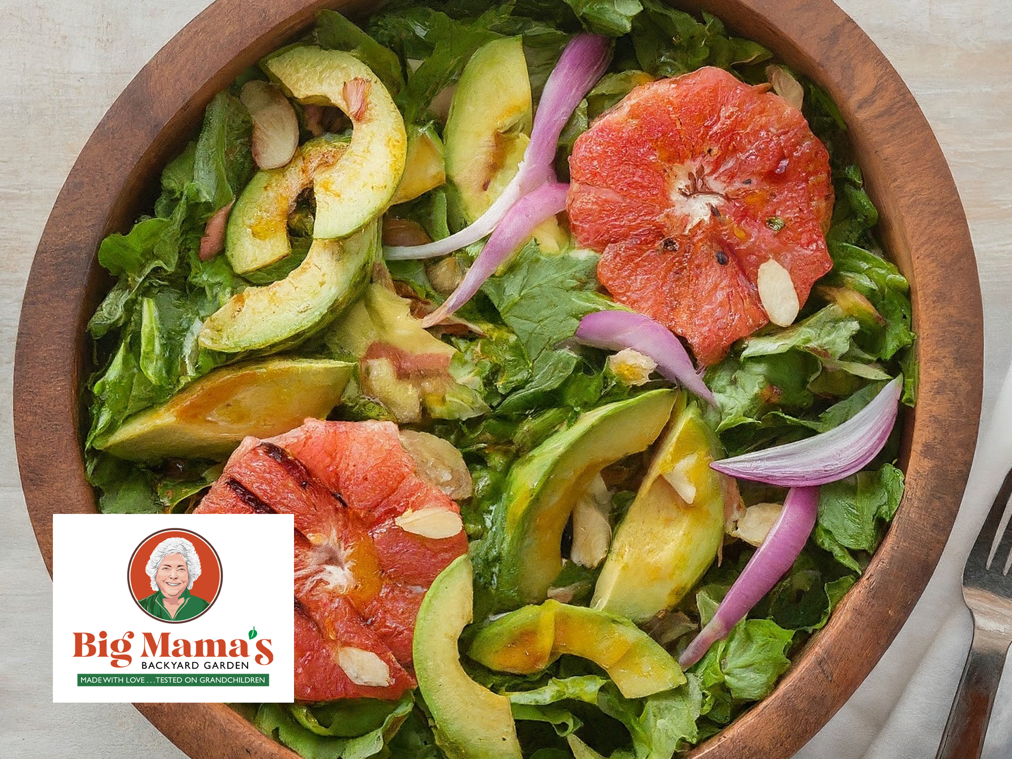 A wooden bowl filled with leafy greens, sliced avocado, red onion, almond slivers, and two large slices of grapefruit makes up this vibrant Citrus Salad. A fork rests beside the bowl and the Big Mama’s Backyard Garden logo appears in the corner.
