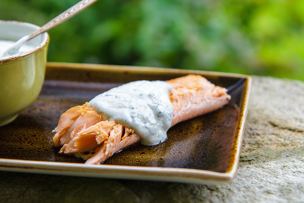 A piece of cooked salmon, made with fresh seafood, is topped with a creamy white dill sauce and served on a brown rectangular plate, with a bowl of sauce and spoon partially visible in the background.