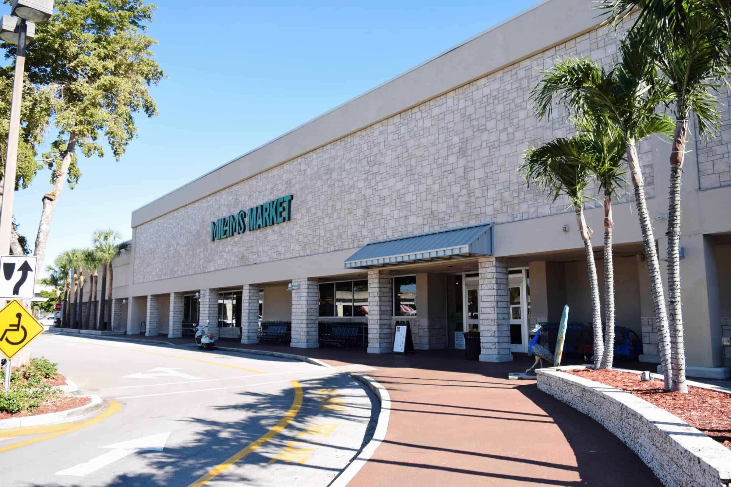 Exterior view of a Milam’s Market grocery store with palm trees, a stone facade, and a sidewalk with a yellow pedestrian crossing sign in the foreground on a sunny day.