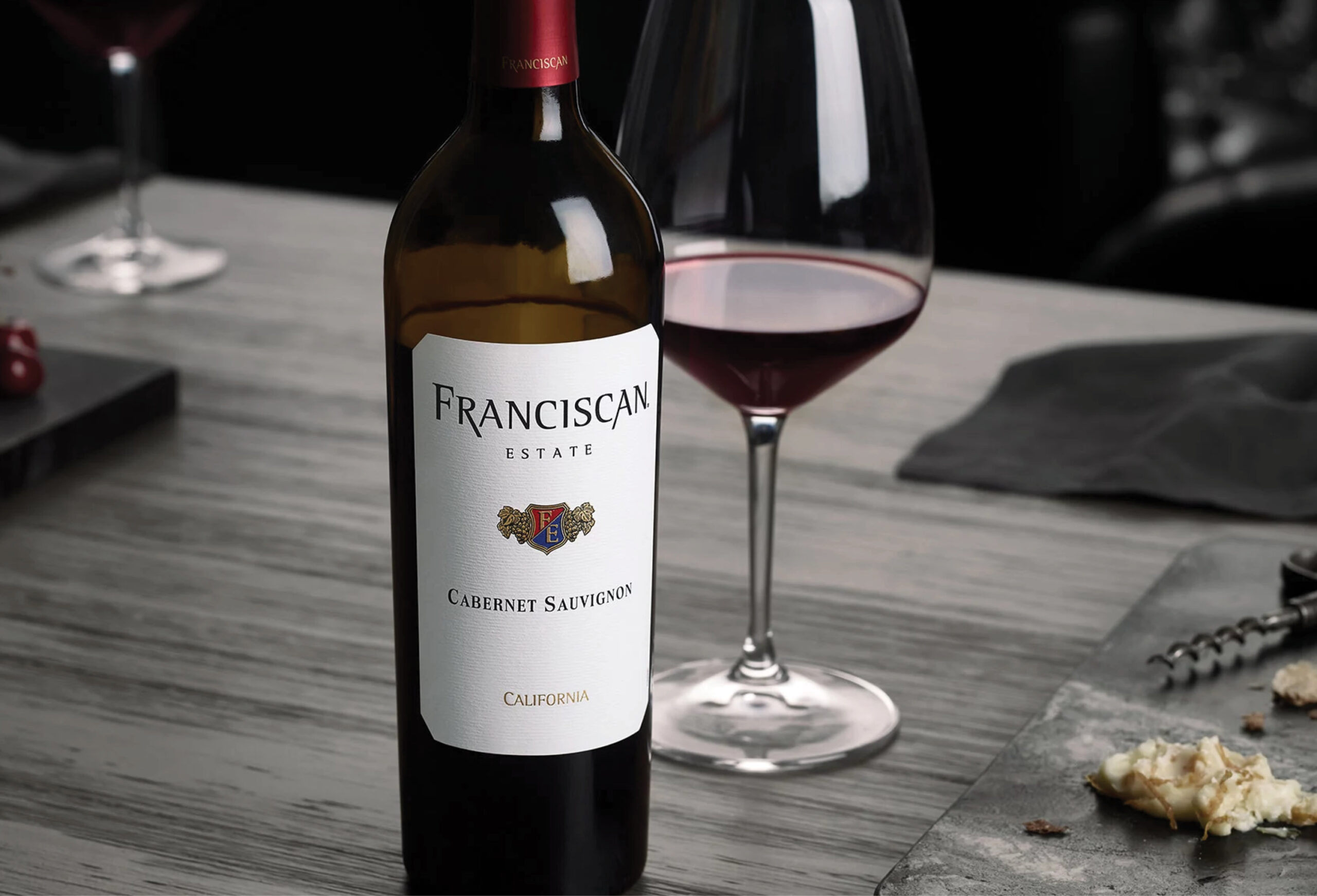 A bottle of Franciscan Estate Cabernet Sauvignon stands on a gray wooden table beside a filled wine glass, a wine opener, and a plate with cheese; the setting is elegant and dimly lit.