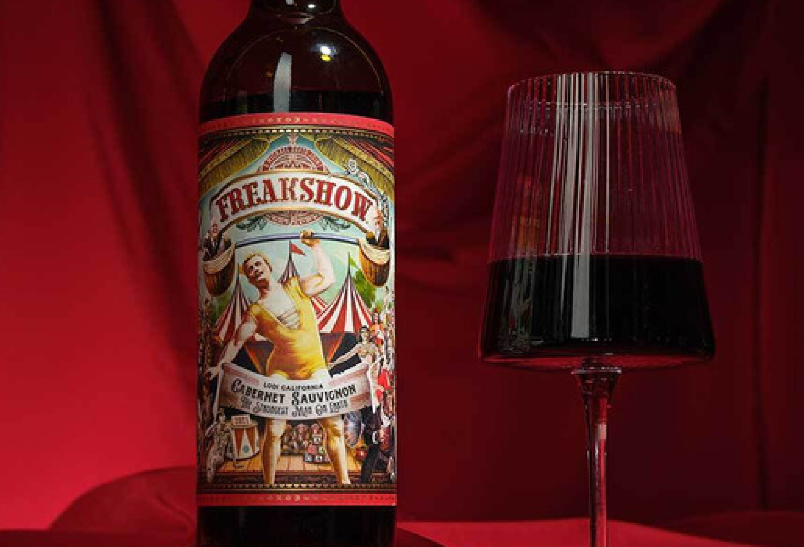 A bottle of Freakshow Cabernet Sauvignon wine stands next to a glass filled with red wine, set against a rich red background. The bottle label features a circus-themed illustration with vibrant colors.