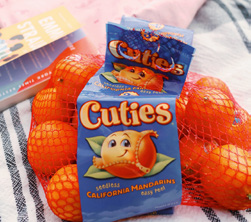 A mesh bag of Cuties seedless California mandarins sits on a striped cloth, with a book partially visible in the background.