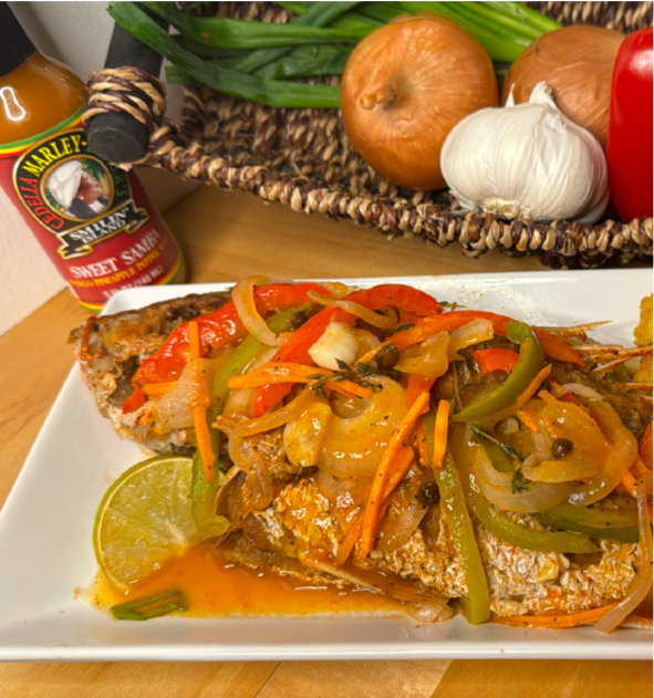 A plated Escovitch Fish, a classic Jamaican fish dish, topped with cooked onions, bell peppers, and carrots in a tangy Sweet Samba sauce, garnished with lime slices. In the background are onions, garlic, scallions, red pepper, and hot sauce.