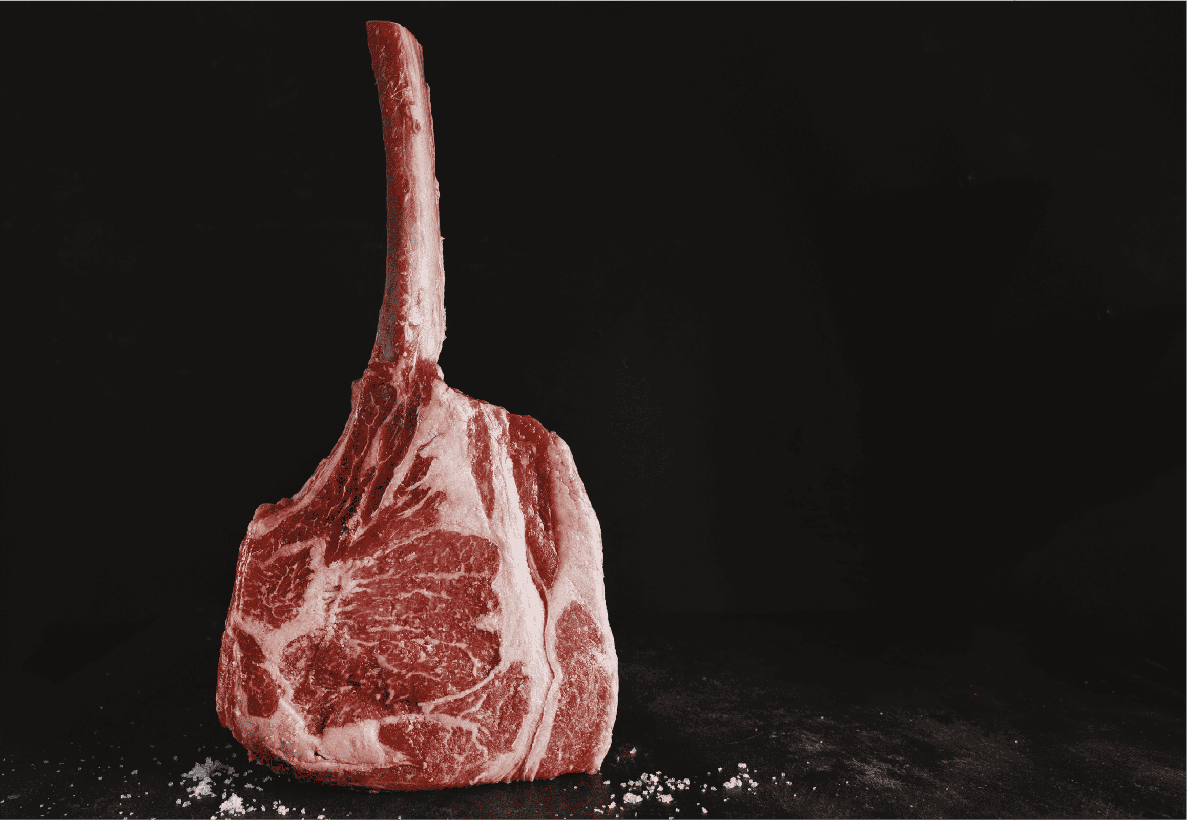 A raw tomahawk steak, one of the most impressive beef cuts, stands upright on a dark surface with rich marbling and coarse salt, set against a black background.