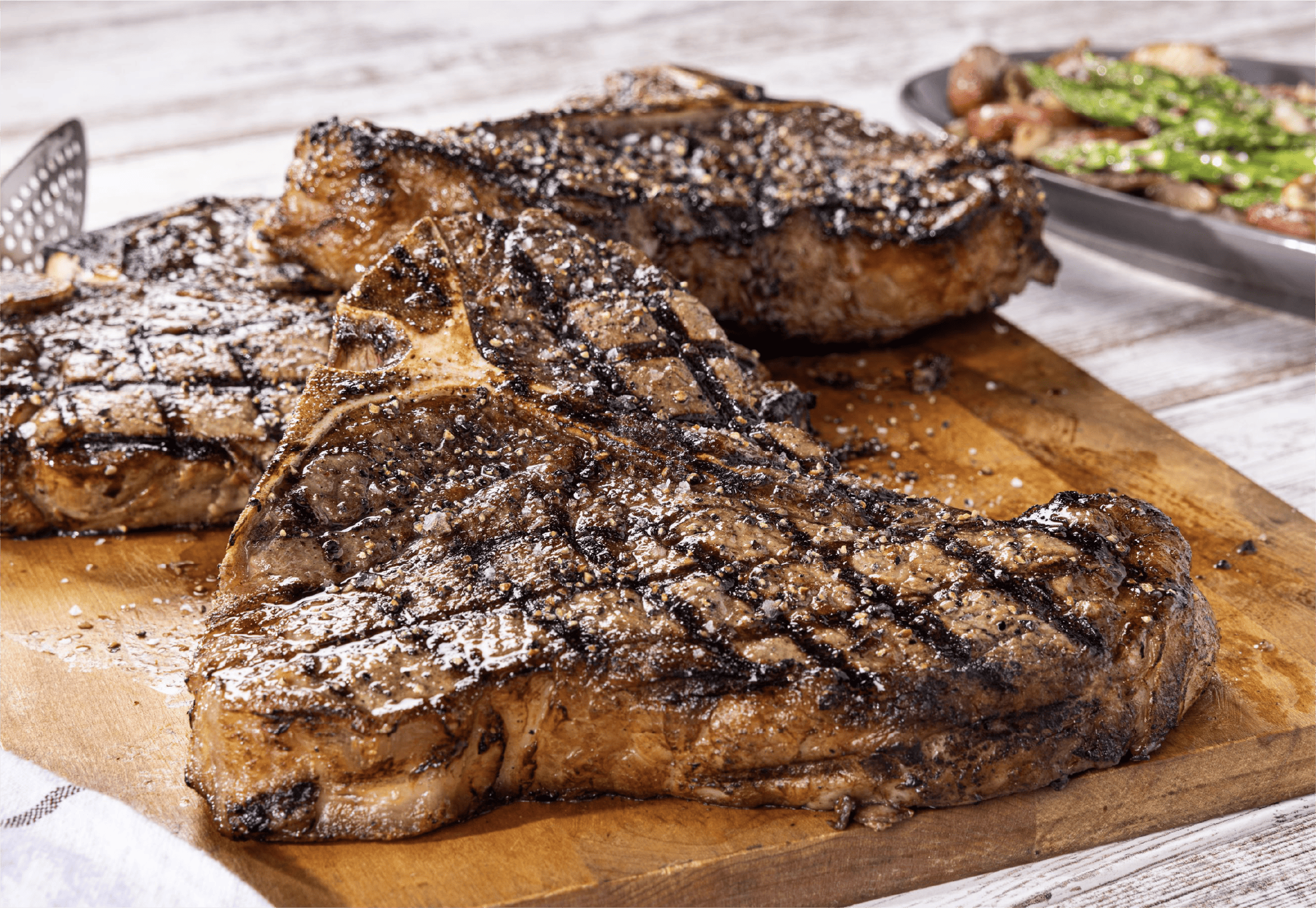 Three grilled T-bone and porterhouse steaks sprinkled with coarse salt rest on a wooden cutting board. Grill marks are visible on the steaks, hinting at a perfect backyard meal, with green vegetables blurred in the background.