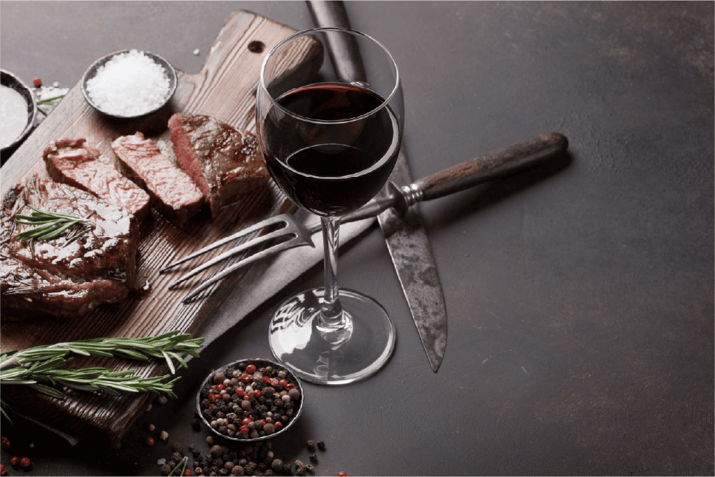 A glass of red wine sits beside sliced steak cuts on a wooden board, garnished with rosemary. A fork, knife, and small bowls of salt and pepper complete the rustic tabletop—perfect for classic wine and steak pairings.