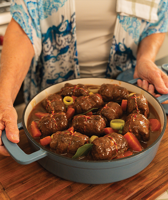 A person holds a blue pot filled with Rouladen, carrots, and leeks in a savory sauce, displayed on a wooden surface—perfect with a side of Rotkohl.