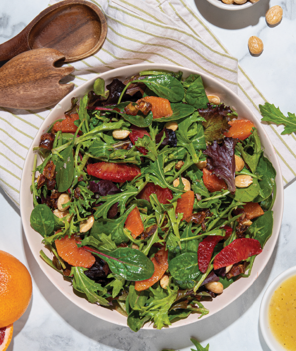 A bowl of Winter Citrus Salad with arugula, spinach, orange and blood orange slices, crunchy almonds, sweet dates, and a light dressing, served on a striped cloth with wooden salad utensils and nuts nearby.