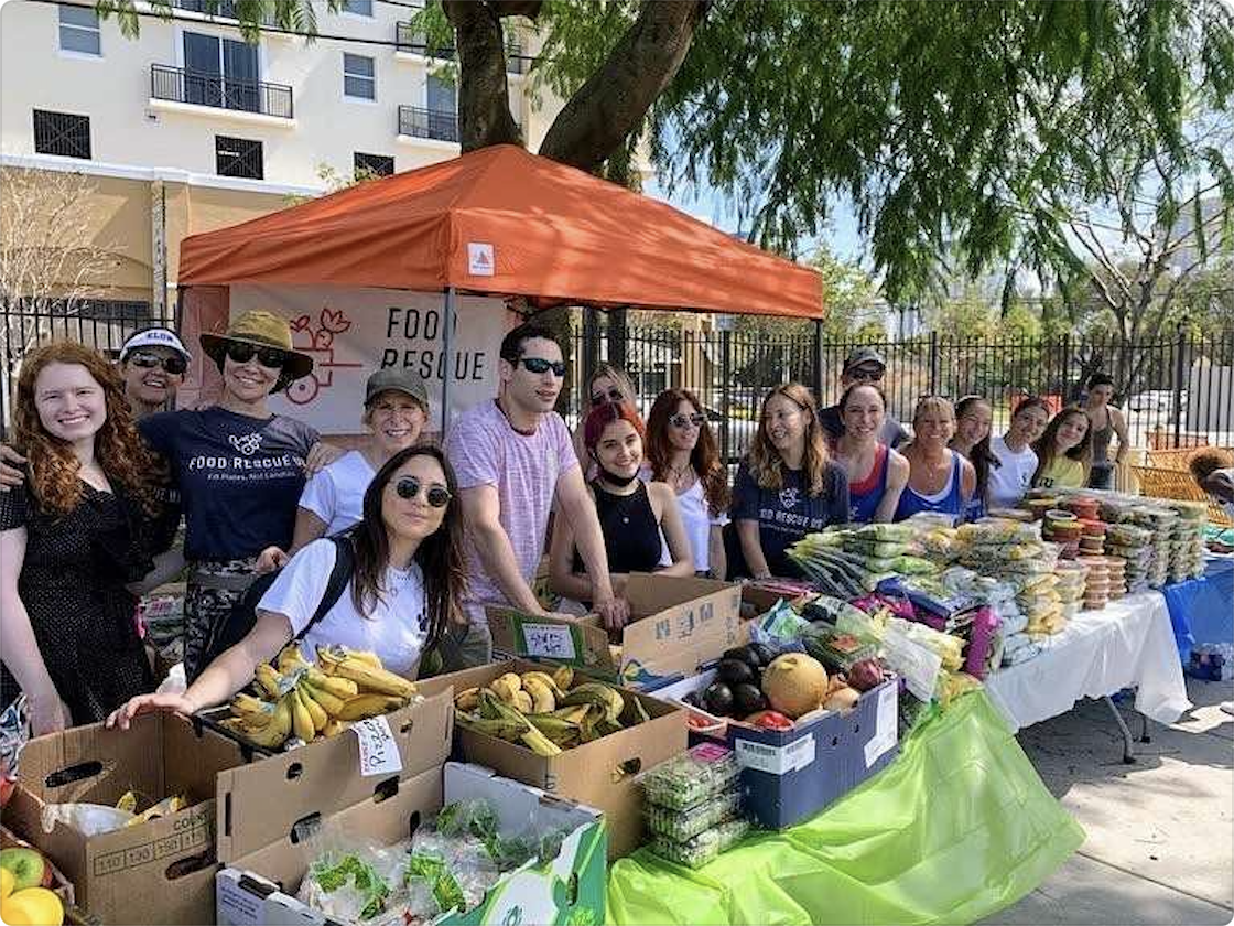 A group of people smiles in front of a table filled with fruits, vegetables, and packaged foods at an outdoor food rescue and fundraising event, under a bright orange canopy with apartment buildings in the background to support local food banks.