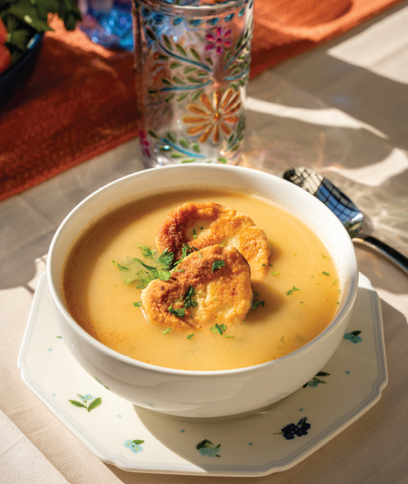 A bowl of creamy Mexican soup, inspired by a traditional recipe, topped with two pieces of toasted bread and chopped herbs, sits on a decorative plate with a glass featuring a colorful floral pattern in the background.