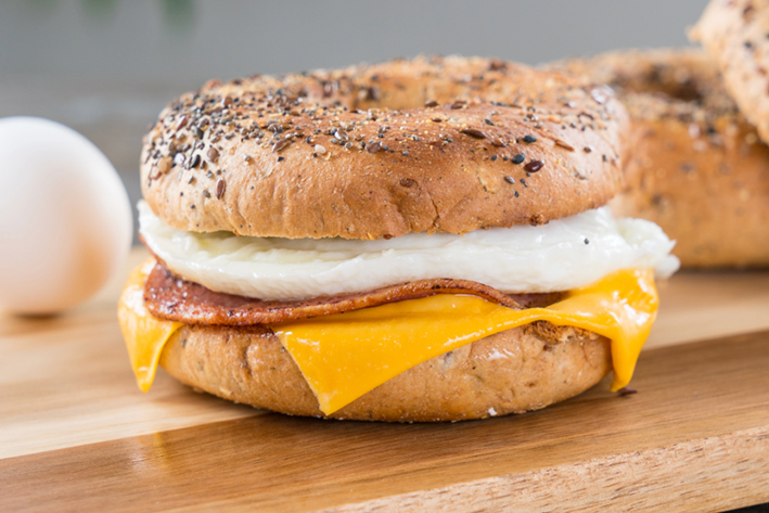 A seeded bagel sandwich with a slice of melted cheddar cheese, a round slice of sausage, and a cooked egg, sitting on a wooden surface. An egg is placed in the background.