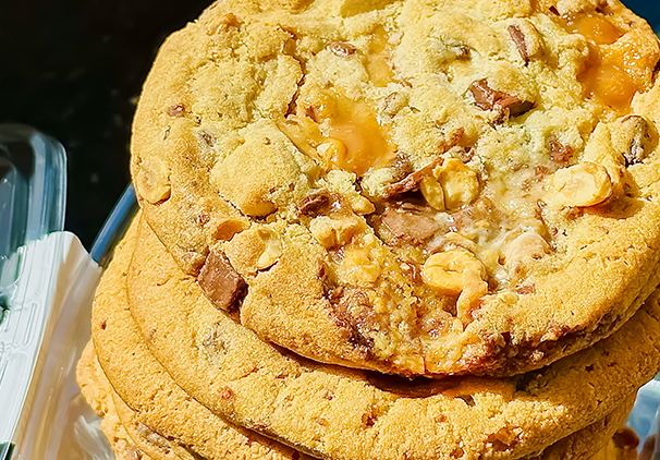 A close-up of a stack of large, golden brown cookies with pieces of nuts and caramel visible, sitting on a clear tray. The cookies look soft and chewy with a slightly crisp edge.