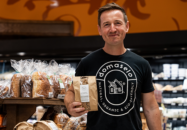 A smiling man wearing a “domaselo” t-shirt holds a bag of bread in a grocery store’s bakery section, with shelves of various loaves in the background.