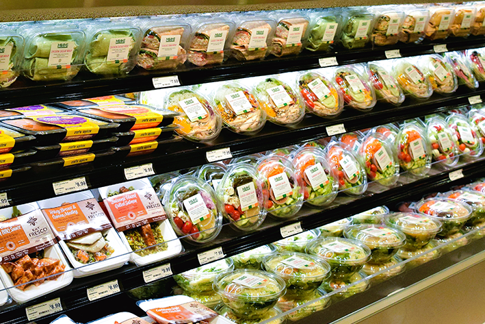 Shelves in a grocery store display a variety of pre-packaged salads and prepared meals in plastic containers, neatly arranged and labeled, ready for purchase.