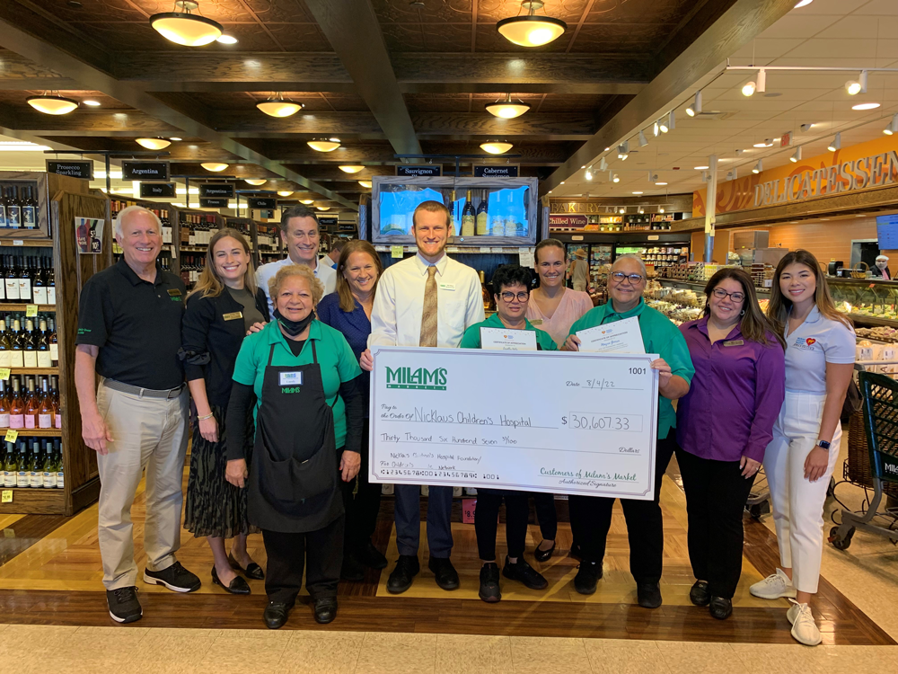 A group of people poses inside a grocery store, holding an oversized check for $30,067.33 made out to Arnold Palmer Children's Hospital, highlighting Community Donations with MIA4MS and Milams Markets visible on the check.