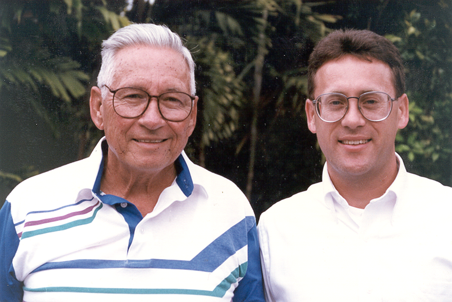 Two men smiling outdoors in front of leafy greenery; one older with white hair and glasses, wearing a striped polo shirt, and one younger with brown hair and glasses, wearing a white button-up shirt.