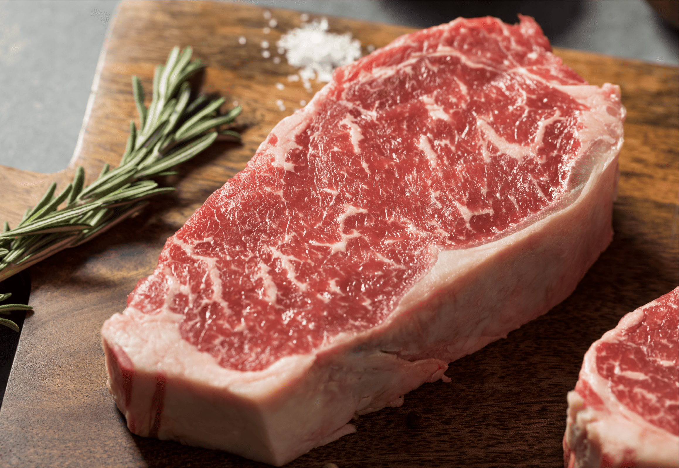 A raw, marbled New York Strip steak on a wooden cutting board, with a sprig of fresh rosemary and a small pile of coarse salt beside it—perfect for exploring Meat Cuts and getting Meat Explained.